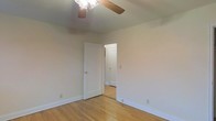 This single-floor space presents a straightforward and functional design. The room visible in the image features light hardwood flooring that adds warmth to the neutral, beige-toned walls. A ceiling fan with wooden blades and an integrated light fixture is mounted overhead, providing both air circulation and ambient lighting. White baseboards frame the perimeter of the room, offering a clean contrast against the flooring. An open doorway connects to an adjacent room or hallway, suggesting a practical, flowing layout between spaces. The arched transition visible beyond the doorway hints at subtle architectural detailing throughout. The overall ambiance is understated and move-in ready, with a simple palette that allows for versatile interior styling.