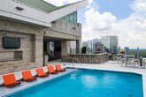 Located in the heart of Atlanta's Buckhead district, this sophisticated amenity floor at 3390 Stratford Rd NE offers a compelling blend of indoor comfort and outdoor recreation. The rooftop pool deck is a standout feature, showcasing a sleek swimming pool flanked by vibrant orange lounge chairs, a stacked-stone fireplace with a wall-mounted TV, and a separate wet bar area — all set against an open Atlanta skyline. Inside, the expansive lounge area is thoughtfully designed with bold teal chevron accent walls, large-format tile flooring transitioning to plush carpet, and ample seating including blue velvet chairs, a wraparound sofa, and ottoman seating. Floor-to-ceiling windows bathe the space in natural light while framing striking panoramic views of the Atlanta skyline. A dedicated conference or private dining room features a substantial dark wood table, upholstered seating, a framed fireplace with a mounted display, and direct glass-door access to the outdoor terrace. The entry corridor presents a clean, modern aesthetic with soft gray-blue walls, tile flooring, curated artwork, and elevator access. Together, these spaces form a polished, high-rise amenity environment ideal for socializing, relaxing, and productive gatherings.
