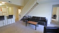 This two-story residence is designed with a practical, open layout that makes efficient use of its space. The main level features a carpeted living area that serves as the central gathering space, furnished with a dark leather sofa and a wooden coffee table with a black frame. A white-painted open-riser staircase with baluster railings is a defining architectural element of the main floor, connecting both levels while adding visual height to the room. Beneath the staircase, a compact shelving unit provides additional storage. Adjacent to the living area, a glimpse of the kitchen or dining space is visible, featuring a bar-style counter with seating, suggesting an open-plan connection between the living and kitchen zones. The neutral, light-toned walls and beige carpeting throughout the main level contribute to a clean, understated ambiance. The layout flows naturally between rooms, offering a cohesive and functional living environment across both floors. The staircase leading to the upper level hints at additional living or sleeping space above, making this a versatile multi-level home.