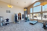 Located in Lakewood, Colorado, this fitness center offers a well-appointed exercise space designed to support a comprehensive workout routine. The room is defined by its dramatic vaulted ceilings, which create an open, airy atmosphere enhanced by a pair of elegant pendant light fixtures. A striking floor-to-ceiling window wall, topped with an arched transom window, floods the space with natural light and provides views of the outdoor pool area beyond.

The exercise room is outfitted with a diverse range of equipment to accommodate various fitness goals. Cardio options include a treadmill, elliptical trainer, stationary bike, and a rowing machine. Strength training is supported by a multi-station cable machine, a dedicated weight bench, a full dumbbell rack, and a selection of medicine balls and resistance accessories. A built-in fireplace with a bold gray accent surround serves as a distinctive architectural focal point, adding warmth and character to the space.

Durable, patterned carpet tiles cover the floor throughout, providing both comfort and practicality. An arched doorway leads to an adjacent room, and multiple entry doors offer convenient access. The overall ambiance is bright, functional, and inviting, making this fitness center a well-equipped amenity for residents of the Lakewood community.
