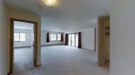 This single-floor space features a generously sized, open living area finished with wall-to-wall light-colored carpeting that lends a clean, neutral aesthetic throughout. The room is illuminated by a central flush-mount ceiling light fixture, while natural light enters through two windows dressed with dark burgundy curtains and a sliding glass door that provides views of the outdoors and access to an exterior area. The sliding door is framed by matching drapery, creating a cohesive look along the exterior wall. Baseboard heating units run along the perimeter, indicating a practical and consistent heating system. The white walls and smooth ceiling contribute to a bright, airy atmosphere. A louvered bifold door is visible along one wall, likely concealing a closet or utility space, while a standard doorway on the opposite side leads to an adjacent room, suggesting a connected floor plan. The overall layout is open and unobstructed, offering flexible use of the main living space. The room's simplicity and neutral finishes provide a versatile foundation suitable for a variety of interior configurations.
