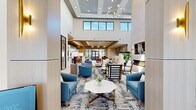 This expansive single-floor amenity space is designed with a contemporary mid-century modern aesthetic, offering a variety of distinct functional areas that flow seamlessly into one another. The grand central lobby commands attention with its soaring double-height ceilings, clerestory windows that flood the interior with natural light, and a striking sputnik chandelier overhead. Patterned area rugs anchor multiple lounge seating arrangements featuring blue upholstered armchairs and marble-topped coffee tables, creating inviting gathering spaces throughout.

A spacious dining and social lounge area showcases light hardwood flooring, a vivid tropical botanical mural as a focal point, and bold cobalt blue cabinetry with gold hardware lining a communal kitchen counter. Comfortable dining tables and chairs are arranged to encourage social interaction, while a fireplace and built-in seating add warmth.

A dedicated screening room features stadium-style seating, floor-to-ceiling drapes, a large wall-mounted display, and an elegant brass chandelier. Private meeting rooms offer intimate round or rectangular tables adorned with curated artwork. A sunlit reading nook with tall windows and curved lounge chairs completes the space, providing serene outdoor views.