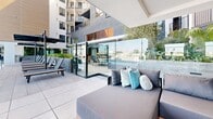 Located at 12888 Crenshaw Blvd in Gardena, California, this rooftop amenity level delivers a comprehensive outdoor living experience designed for relaxation and recreation. The centerpiece is a large, resort-style swimming pool finished in bright aquamarine tile, surrounded by light-toned porcelain pavers and modern wood-slatted chaise lounges that invite sunbathing beneath the Southern California sky. Expansive, unobstructed panoramic views of the surrounding Gardena cityscape and distant mountains define the outdoor atmosphere.

Adjacent to the pool, a semi-enclosed spa room features an indoor hydrotherapy pool framed by textured stone-tiled walls, large windows that admit ample natural light, and lush potted greenery that adds warmth to the space.

A covered lounge and recreation area extends along the rooftop perimeter, featuring polished terrazzo-style flooring, a full-sized ping-pong table, built-in planter beds with drought-tolerant landscaping, and comfortable modular seating arranged around a wall-mounted television and fireplace. Floor-to-ceiling glass railings and open-air sightlines maintain a seamless connection between the indoor and outdoor zones. The overall design is distinctly contemporary, combining clean lines, neutral finishes, and abundant greenery to create a refined, resort-inspired community amenity space.