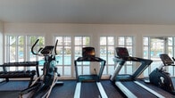 This well-appointed fitness center at 820 Macmillan Avenue in Wilmington, NC offers a comprehensive workout facility designed to accommodate a wide range of exercise routines. The single-floor space is bathed in natural light, courtesy of a generous row of large windows along one wall that frame views of the outdoor swimming pool, creating an open and energizing atmosphere.

The cardio section is outfitted with multiple Promaxima treadmills and elliptical machines positioned directly in front of the pool-facing windows, providing an inspiring backdrop during workouts. A dedicated free weight area features a fully stocked dumbbell rack with a wide range of weights, ideal for strength training. The space also includes cable and multi-function strength machines, a recumbent bike, a spin bike, and a leg press machine, offering a thorough full-body workout experience.

For flexibility and functional training, stability balls and resistance equipment are available in a designated area accompanied by a wall-mounted flat-screen TV. Additional TVs are placed throughout the space for entertainment during workouts. The flooring consists of durable patterned carpet throughout, while the vaulted ceiling with exposed ductwork and recessed lighting adds an industrial-modern touch. Convenient amenities include a water fountain and hand sanitizer stations.