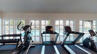 This well-appointed fitness center at 820 Macmillan Avenue in Wilmington, NC offers a comprehensive workout facility designed to accommodate a wide range of exercise routines. The single-floor space is bathed in natural light, courtesy of a generous row of large windows along one wall that frame views of the outdoor swimming pool, creating an open and energizing atmosphere.

The cardio section is outfitted with multiple Promaxima treadmills and elliptical machines positioned directly in front of the pool-facing windows, providing an inspiring backdrop during workouts. A dedicated free weight area features a fully stocked dumbbell rack with a wide range of weights, ideal for strength training. The space also includes cable and multi-function strength machines, a recumbent bike, a spin bike, and a leg press machine, offering a thorough full-body workout experience.

For flexibility and functional training, stability balls and resistance equipment are available in a designated area accompanied by a wall-mounted flat-screen TV. Additional TVs are placed throughout the space for entertainment during workouts. The flooring consists of durable patterned carpet throughout, while the vaulted ceiling with exposed ductwork and recessed lighting adds an industrial-modern touch. Convenient amenities include a water fountain and hand sanitizer stations.