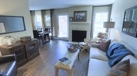 This single-floor residence offers a thoughtfully designed open-concept layout that seamlessly connects the living and dining areas into one cohesive, welcoming space. Rich, dark hardwood flooring runs throughout, lending warmth and a sense of continuity to the interior. The living area is anchored by a classic fireplace with a light-toned stone surround and a simple white mantel, creating a natural focal point for the room. Above the mantel, a framed artwork and a decorative clock add character and a personalized touch. The space is generously filled with natural light, drawn in through multiple large windows dressed with neutral curtains and adjustable blinds, as well as a glass-paned door that opens to an outdoor area, reinforcing a bright and airy atmosphere. The dining area, positioned adjacent to the living space, accommodates a rustic wooden dining table and chairs, perfectly complementing the warm tones of the flooring. A media console along one wall provides functional storage while enhancing the room's traditional aesthetic. Comfortable seating, including a sofa and accent chairs with decorative pillows, complete the living area. Overall, the space presents a harmonious blend of classic design elements and practical living, offering a comfortable and well-lit environment throughout.