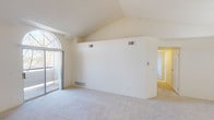 This single-floor unit offers a thoughtfully designed layout that maximizes both openness and functionality. The main living area immediately impresses with its dramatic vaulted ceiling that rises to a sharp peak, creating a strong sense of vertical space and airiness. A large arched window paired with a sliding glass door floods the room with natural light and provides views of the surrounding outdoor greenery. Soft wall-to-wall carpeting runs throughout the living, dining, and bedroom areas, contributing to a warm and comfortable atmosphere.

The kitchen is positioned in an open-concept arrangement, separated from the living space by a white peninsula that offers additional workspace and serves as a casual dining counter. The kitchen is equipped with white appliances including a refrigerator, range, and microwave, complemented by wood-trimmed cabinetry that adds a touch of warmth to the otherwise light and neutral palette.

The bedroom is a calm and private retreat, featuring plush carpeting, neutral walls, and a window that brings in soft natural light while framing views of the outdoors. The bathroom is clean and functional, featuring a full tub and shower combination with white tile surround accented by a distinctive teal tile border, adding a subtle decorative detail to the space. Overall, the unit presents a clean, light-filled environment with consistent neutral tones throughout.