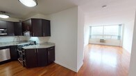 This single-floor unit offers a well-organized layout with distinct living, kitchen, and bathroom areas, all unified by warm-toned hardwood flooring throughout. The open living area is a standout feature, benefiting from a large picture window that floods the space with natural light and provides an airy, open feel. A wall-mounted HVAC unit ensures year-round climate control.

The kitchen is efficiently designed in an L-shaped configuration, featuring rich dark espresso cabinetry that extends to the ceiling for ample storage. Granite-look countertops provide a clean, modern aesthetic, while a full suite of stainless steel appliances — including a refrigerator, dishwasher, microwave, and gas range — offers practical functionality. The kitchen opens toward the living area, separated by a partial wall that adds architectural definition without fully closing off the space.

The bathroom is finished in a crisp, all-white palette, with mosaic tile lining the tub surround and shower walls. Brushed nickel fixtures, a wall-mounted sink, and a recessed mirror cabinet contribute to a clean, contemporary design. White walls throughout the unit provide a neutral backdrop, making the space feel fresh and move-in ready.