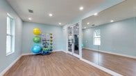 This purpose-built yoga and fitness room offers a clean, calming environment designed to support a variety of wellness activities. The walls are painted in a soft, pale blue tone that creates a tranquil atmosphere conducive to yoga, stretching, and light exercise. Smooth hardwood-style flooring runs throughout the space, providing a practical yet attractive surface suitable for bare-foot workouts and yoga practice.

Recessed lighting is evenly distributed across the white ceiling, ensuring the room is well-lit without harsh glare. A large wall-to-wall mirror on one side of the room enhances the sense of space and allows users to monitor their form during exercise. A window on the adjacent wall introduces natural light, adding to the airy, open feel of the room.

A freestanding equipment rack near the corner keeps the space organized, housing an assortment of exercise balls in various sizes and colors, resistance bands, and additional fitness accessories, making everything readily accessible. French doors open directly into a separate, fully equipped gym area, providing a seamless connection between the two fitness spaces for a comprehensive workout experience.