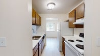 This single-floor unit offers a well-organized layout designed for functional everyday living. The galley-style kitchen is lined on both sides with warm wood-toned cabinetry, providing generous upper and lower storage. White appliances — including a coil-burner electric range, refrigerator, and dishwasher — are paired with white laminate countertops and a stainless steel sink. Light wood-look flooring runs throughout the kitchen, which opens at the far end into the living area through a pass-through counter, creating a connected feel between the two spaces.

The living area is carpeted and open, receiving natural light from a window on the side wall. The kitchen pass-through adds visual connectivity and a casual bar-style interaction with the space beyond.

The bedrooms are carpeted and neutrally finished, each featuring windows dressed with vertical blinds that allow outdoor views and natural light. One bedroom includes a walk-in closet with built-in shelving, while another features a standard closet. The bathroom is clean and simply appointed, with white tile surrounding a full soaking tub and shower combination, light wood-look flooring, and brushed nickel fixtures and grab bar hardware throughout.