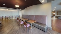 This thoughtfully designed communal space combines lounge, dining, and kitchen functions within a single open-plan layout. A striking architectural detail defines the interior: a curved, dark-painted ceiling panel that contrasts with the surrounding light-colored walls, adding visual depth and a modern, clubhouse-quality ambiance throughout.

At the heart of the space sits a large, curved kitchen island topped with granite-style countertops and lined with round-seated bar stools. The adjacent kitchenette wall features dark cabinetry, a stainless steel refrigerator, a wall-mounted television, and a built-in microwave, making the area well-suited for both casual gatherings and catered events. A globe chandelier overhead adds a warm, decorative touch.

The dining and lounge area is anchored by an extended upholstered banquette finished in a silver-and-purple striped fabric, paired with warm wood-topped tables and coordinating pink-upholstered chairs. High-top tables and seating near large windows allow natural light to fill the space, offering views of an adjacent landscaped courtyard.

Glass doors with full-height windows open directly to an outdoor patio, seamlessly extending the gathering space outside. Wood-look flooring unifies the entire layout, contributing to the space's warm and inviting character.