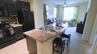 This single-floor residence showcases an open-concept layout that effortlessly connects the kitchen and living areas into one cohesive, functional space. The kitchen is appointed with a curved granite countertop island that doubles as a breakfast bar, complete with seating for two bar stools and an integrated sink with a stainless steel faucet. Black appliances, including a full-size refrigerator, lend a bold, modern contrast to the lighter tones of the cabinetry and countertops. Wood-look flooring anchors the kitchen area, adding warmth and easy maintenance. The open design transitions naturally into the carpeted living room, which is bathed in natural light streaming through multiple windows dressed with green curtain panels. A ceiling fan with integrated lighting keeps the space comfortable and well-lit. The living area is generously proportioned, offering room for a full sofa and accent seating, with neutral wall tones providing a versatile backdrop. The overall ambiance is bright, airy, and welcoming, making it an ideal space for both everyday living and casual entertaining.