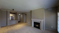 This single-floor residence offers a thoughtfully designed layout with a comfortable, cohesive aesthetic anchored by warm neutral tones and soft carpeted flooring throughout the main living area. The living room serves as the focal point of the space, highlighted by a classic white-mantled fireplace with a tiled surround — a standout architectural detail that adds both warmth and character to the room. Crown molding traces the ceiling, lending a polished, refined finish to the space.

Open sightlines connect the living area to the kitchen, which features a raised bar-style counter with granite or stone countertops, providing a natural transition between cooking and living spaces. White cabinetry in the kitchen contributes to a clean, bright contrast against the warmer tones of the surrounding walls.

A hallway visible from the living room leads to additional areas of the residence, including what appears to be a closet and further private rooms, suggesting a well-organized and functional floor plan. Recessed and flush-mount lighting provides consistent illumination throughout. The overall ambiance is calm and inviting, with a neutral palette of taupe and beige tones creating a versatile backdrop suited to a variety of interior styles.