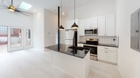 This single-floor space in Greenville, SC, presents a clean, contemporary aesthetic throughout. The kitchen serves as a central highlight, showcasing white shaker cabinetry, dark granite countertops, and a coordinating island with a beadboard-paneled base. Stainless steel appliances—including a refrigerator, range, and built-in microwave—complement the subway tile backsplash, while a pair of matte black pendant lights hang above the island, adding a bold design contrast. Light wood-tone flooring flows seamlessly throughout the space, enhancing the bright, airy feel created by the crisp white walls.

The living area is open and versatile, featuring recessed lighting and a ceiling fan, offering a blank canvas for customization. A separate bedroom benefits from natural light through a large black-framed window that contrasts against the exposed brick exterior wall visible outside, lending an industrial character to the space.

The bathroom features a walk-in shower finished with floor-to-ceiling horizontal white tile, a matte black shower fixture, a built-in niche, and a classic penny-tile floor. The consistent use of matte black accents and white tones throughout ties the design together cohesively.