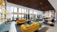 This thoughtfully designed single-floor space makes an immediate impression with its dramatic double-height ceilings and expansive floor-to-ceiling windows that flood the interior with natural light and offer open views of the surrounding outdoors. The architectural design blends warm wood-slatted ceiling panels with crisp white walls, creating a refined contrast that feels both modern and inviting.

The open-plan lounge area is generously proportioned, providing ample space for relaxed gathering and socializing. Bold mustard-yellow sofas are arranged around low glass coffee tables, anchored by a large area rug in cool slate tones. Complementing these are teal upholstered benches and patterned accent chairs, adding depth and visual interest to the color palette.

Pendant light fixtures hang elegantly from the high ceiling, contributing to the warm ambiance alongside recessed lighting throughout. Toward the rear, a built-in shelving unit and a reception or service counter area extend the functionality of the space. Lush potted plants are strategically placed to introduce natural elements.

The abundance of glazing on multiple walls ensures the space feels airy and connected to the exterior environment at all times, making it an engaging and comfortable destination for occupants throughout the day.