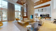 This expansive single-floor amenity space is designed with a contemporary aesthetic that seamlessly blends comfort and functionality. The centerpiece of the main lounge is a dramatic floor-to-ceiling stacked stone fireplace flanked by a striking geometric wood-framed decorative screen, anchoring the double-height volume of the space. Warm wood-paneled ceiling insets contrast beautifully with crisp white walls, while large-format tile flooring runs throughout the open layout.

Generous floor-to-ceiling windows and glass doors flood the interior with natural light and offer views of an outdoor courtyard area. The lounge is furnished with neutral upholstered seating, accent chairs in mustard yellow, and marble-topped side tables that introduce gold-toned metallic accents throughout.

A dedicated game area accommodates both a ping-pong table and a billiards table, providing recreational appeal. Adjacent to the main lounge, a more intimate seating area features a warm wood ceiling, additional soft seating, and a bold diamond-patterned display wall. A sleek kitchenette bar lined with dark glazed tile and under-cabinet lighting offers beverage and refreshment service. The overall atmosphere is polished, welcoming, and well-appointed.