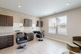 Located at 6565 Chupp Rd in Stonecrest, GA, this single-floor salon space is thoughtfully designed to deliver a polished and functional grooming experience. The main styling area features two professional barber/styling chairs, each positioned at its own dark-espresso cabinetry station complete with granite-topped counters, integrated shampoo bowls, and upper storage cabinets. A striking hexagonal tile accent wall serves as the visual centerpiece of the room, lending a modern, upscale aesthetic that contrasts elegantly with the neutral beige walls and large-format light tile flooring. Recessed lighting overhead ensures bright, even illumination throughout the space. A generously sized window fitted with horizontal blinds allows ample natural light to filter in while offering views of the surrounding community. Large mirrors are mounted above each station, enhancing the sense of openness within the compact footprint. A separate waiting area provides a welcoming transition into the salon, furnished with a plush sage-green accent chair and ottoman beneath a colorful framed landscape print. The space is clean, contemporary, and efficiently organized, making it well-suited for professional hair and grooming services in a comfortable, boutique-style setting.