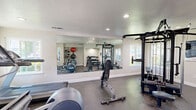 Located at 3825 Little Rock Drive in Antelope, California, this fitness center offers a well-equipped exercise space designed to support a full range of workout routines. The room features a textured ceiling with recessed lighting that provides bright, even illumination throughout. Durable speckled rubber flooring covers the entire area, offering both safety and comfort underfoot.

The cardio section is outfitted with Precor equipment, including an elliptical trainer, a stationary upright bike, and a treadmill, all positioned near a window that allows in natural light. A wall-mounted flat-screen TV is conveniently placed for viewing during workouts. On the strength training side, a multi-station cable machine serves as the centerpiece, complemented by an adjustable weight bench. A three-tier dumbbell rack holds a full set of free weights, while a dedicated wall-mounted rack holds exercise balls and resistance bands for functional training.

A large floor-to-ceiling mirror along one wall visually expands the space and allows users to monitor their form. Windows on multiple walls bring in natural light and views of the surrounding outdoor greenery. The entrance features a glass-panel door, a posted fitness center rules sign, and a security camera, ensuring a safe and organized environment. The overall ambiance is clean, functional, and inviting.