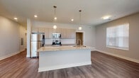 Located at 2875 Olentangy River Rd in Columbus, Ohio, this single-floor residence offers a thoughtfully designed open-concept layout with modern finishes throughout. The kitchen serves as the heart of the home, showcasing a large granite-topped island with an integrated sink, globe pendant lighting, flat-front cabinetry in a warm taupe finish, a white subway tile backsplash, and a full suite of stainless steel appliances including a refrigerator, range, and over-the-range microwave. The kitchen flows seamlessly into the living and dining areas, which are bathed in natural light from multiple windows fitted with horizontal blinds. Rich wood-look plank flooring spans the main living areas, adding warmth and continuity to the space. The primary bathroom features a granite-topped vanity with a framed mirror, stylish lighting fixtures, and a full tub-and-shower combination dressed in elegant marble-style tile surround. A generously sized walk-in closet with wire shelving provides ample storage. The bedroom is carpeted for comfort and includes a window with natural light and outdoor views. A second bathroom mirrors the same quality finishes, complete with a granite vanity, framed mirror, and a separate shower. The overall aesthetic is clean, contemporary, and move-in ready.