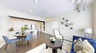 This single-floor residence showcases a thoughtfully designed open-concept layout that seamlessly connects the living, dining, and kitchen areas into one cohesive and functional space. Dark hardwood floors run throughout, providing a striking contrast against the light, neutral-toned walls and white cabinetry. Recessed lighting and track fixtures illuminate the interior, creating a bright and welcoming ambiance.

The kitchen is well-appointed with stainless steel appliances, including a refrigerator and microwave, and features a peninsula with bar seating — ideal for casual dining or entertaining. The adjacent dining area accommodates a full table with comfortable seating, anchored by lush greenery that adds a natural touch to the space.

The living area is generously proportioned, offering room for a full seating arrangement centered around a dark round coffee table and complemented by a patterned area rug. A decorative wall display of circular mirrors adds an artistic focal point to the room. Clean white walls, tall ceilings, and an open flow between rooms contribute to an airy, modern atmosphere. The layout is practical and polished, making it well-suited for both everyday living and hosting guests.