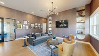 This well-appointed leasing office in Hendersonville, NC offers a welcoming, open-plan environment that seamlessly blends professional functionality with residential comfort. The space is characterized by soaring ceilings, light wood-look tile flooring, and warm, textured wall paneling in soft taupe tones that lend a refined, contemporary feel throughout.

The lounge area anchors one side of the space, featuring a plush gray sectional sofa, amber leather accent chairs, and a rounded natural wood coffee table arranged atop a layered blue area rug. A wall-mounted flat-screen television, built-in arched shelving with curated décor, and a striking sputnik-style chandelier with exposed bulbs add visual interest and personality. A gallery wall of framed artwork and prints further enhances the inviting atmosphere.

Transitioning toward the leasing desk area, a substantial island-style workstation with a crisp white countertop and dark cabinetry serves as the functional hub of the office. Cascading pendant globe lights illuminate the desk from above, while additional wall art and decorative accents maintain a cohesive, curated aesthetic. Large glass doors and windows allow abundant natural light to flood the interior, offering views of the surrounding greenery and ensuring the space feels bright and open throughout the day.