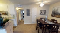 This single-floor residence offers a practical and comfortable layout designed for everyday living. The dining area is a central feature, accommodating a raised bar-height table with cushioned seating, ideal for casual meals or entertaining. Neutral-toned carpet flows through the main living spaces, complemented by warm beige walls that create a cohesive and inviting atmosphere throughout.

A ceiling fan with integrated lighting fixture serves the dining area, providing both air circulation and overhead illumination. The space is well-ventilated with visible HVAC vents positioned throughout the ceiling and walls. Decorative wall art adds personality to the dining space, enhancing the overall aesthetic.

A hallway connects the main living and dining areas to the bedroom, which features its own carpeted flooring and access to additional closet space. The kitchen is positioned adjacent to the dining area, offering a seamless transition between cooking and dining spaces, and is equipped with white cabinetry and appliances. A textured ceiling adds a classic finish detail visible throughout the unit. A utility closet door is also accessible from the dining area, adding practical storage to the layout. The overall design prioritizes functionality while maintaining a warm and welcoming residential feel.