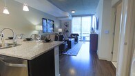 This single-floor space showcases a thoughtfully designed open-concept layout that seamlessly connects the kitchen, dining, and living areas. Rich hardwood flooring runs throughout, adding warmth and elegance to the contemporary aesthetic. The kitchen features a granite-topped island with bar seating and dark cabinetry, providing both a functional workspace and a casual dining area. The living room is comfortably appointed with stylish furnishings, a patterned area rug, and wall art that adds character to the space. A wall-mounted flat-screen television anchors one end of the room, while floor-to-ceiling sliding glass doors at the far end flood the interior with natural light and open onto a private balcony, offering views of the surrounding urban landscape. The tray ceiling detail adds an architectural element that enhances the sense of depth and dimension overhead. The overall ambiance is modern and polished, blending functional design with upscale finishes to create a cohesive and inviting living environment suited for both everyday living and entertaining.