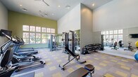 This fitness center in Arcadia Lakes, SC, offers a thoughtfully designed exercise space that accommodates a wide range of workout needs. The single-room facility is characterized by soaring vaulted ceilings that create an open, airy atmosphere, enhanced by large multi-pane windows along the walls that flood the space with natural light and provide views of the surrounding outdoors. Ceiling fans overhead contribute to a comfortable environment during workouts.

The floor is finished with a distinctive patterned carpet tile in neutral gray and yellow tones, providing both durability and visual appeal. Full-length mirrors line one wall, amplifying the sense of space and allowing users to monitor their form. The walls are painted in a crisp white and sage green color scheme, lending the room a clean, energizing aesthetic.

Equipment is well-organized throughout the space and includes multiple treadmills positioned near the windows, elliptical trainers, a rowing machine, and a cable/multi-station strength machine. A dedicated free weights area features a fully stocked dumbbell rack alongside kettlebells and medicine balls for functional training. Adjustable benches and resistance accessories round out the strength training offerings. Wall-mounted televisions are positioned for easy viewing from the cardio equipment, enhancing the workout experience.