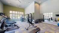 This fitness center in Arcadia Lakes, SC, offers a thoughtfully designed exercise space that accommodates a wide range of workout needs. The single-room facility is characterized by soaring vaulted ceilings that create an open, airy atmosphere, enhanced by large multi-pane windows along the walls that flood the space with natural light and provide views of the surrounding outdoors. Ceiling fans overhead contribute to a comfortable environment during workouts.

The floor is finished with a distinctive patterned carpet tile in neutral gray and yellow tones, providing both durability and visual appeal. Full-length mirrors line one wall, amplifying the sense of space and allowing users to monitor their form. The walls are painted in a crisp white and sage green color scheme, lending the room a clean, energizing aesthetic.

Equipment is well-organized throughout the space and includes multiple treadmills positioned near the windows, elliptical trainers, a rowing machine, and a cable/multi-station strength machine. A dedicated free weights area features a fully stocked dumbbell rack alongside kettlebells and medicine balls for functional training. Adjustable benches and resistance accessories round out the strength training offerings. Wall-mounted televisions are positioned for easy viewing from the cardio equipment, enhancing the workout experience.