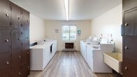 Located at 2227 W Willow Knolls Dr in Peoria, Illinois, this single-floor community facility offers a range of shared amenities designed to support everyday living. The laundry room is generously sized, lined on both sides with rows of top-loading washers and dryers finished in white. Light wood-look flooring runs throughout, and a window with horizontal blinds allows natural light to filter in, creating a bright and functional workspace. A wall-mounted unit provides additional climate control for comfort during use. Adjacent to the laundry area is a dedicated storage and folding zone, featuring a long laminate countertop ideal for sorting and folding laundry, alongside a wall of dark metal lockers with individual locking compartments — offering secure personal storage for residents or users of the facility. A bulletin board is mounted nearby for community notices. The common lounge area provides a welcoming, relaxed setting with carpeted flooring, a full-size sofa, upholstered armchair, glass-top side table, and a large work or dining table with seating. Decorative wall accents give the space a homey character, while overhead fluorescent lighting ensures the room remains well-lit. Together, these spaces form a practical and community-oriented facility suited for shared living or transitional housing environments.