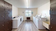 Located at 2227 W Willow Knolls Dr in Peoria, Illinois, this single-floor community facility offers a range of shared amenities designed to support everyday living. The laundry room is generously sized, lined on both sides with rows of top-loading washers and dryers finished in white. Light wood-look flooring runs throughout, and a window with horizontal blinds allows natural light to filter in, creating a bright and functional workspace. A wall-mounted unit provides additional climate control for comfort during use. Adjacent to the laundry area is a dedicated storage and folding zone, featuring a long laminate countertop ideal for sorting and folding laundry, alongside a wall of dark metal lockers with individual locking compartments — offering secure personal storage for residents or users of the facility. A bulletin board is mounted nearby for community notices. The common lounge area provides a welcoming, relaxed setting with carpeted flooring, a full-size sofa, upholstered armchair, glass-top side table, and a large work or dining table with seating. Decorative wall accents give the space a homey character, while overhead fluorescent lighting ensures the room remains well-lit. Together, these spaces form a practical and community-oriented facility suited for shared living or transitional housing environments.