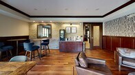 Located in the heart of Saint Paul, MN, this stylish single-floor lounge and gathering space blends classic architectural character with modern interior design. Warm hardwood floors run throughout, complemented by exposed yellow brick walls, dark wood crown molding, and wainscoting that lend a refined, club-like atmosphere.

The space is organized into distinct seating zones that serve a variety of uses. A casual café-style area features high-top tables with bar stools positioned near a beverage station with rich mahogany cabinetry — ideal for a quick coffee or informal meeting. Adjacent to this, a sunlit seating nook with large picture windows offers sweeping street-level views of the surrounding neighborhood, including a striking historic church facade, and is furnished with contemporary blue upholstered chairs and rustic wood tables.

A reading lounge area adds an intimate dimension, featuring leather armchairs, open metal shelving with curated décor, and nature-inspired wallpaper depicting a birch tree motif. A dramatic drum pendant light anchors the space.

The main lounge area is expansive and light-filled, surrounded by floor-to-ceiling windows that frame views of a lush urban park. Deep leather sofas and chairs are arranged around a textured round coffee table, creating an inviting and comfortable communal retreat. The overall ambiance is warm, sophisticated, and thoughtfully designed.