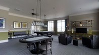 This single-floor game room is thoughtfully designed for recreation and social gatherings. The space is anchored by two standout gaming features: an oval poker table with cushioned chairs positioned in the foreground, illuminated by a striking rectangular pendant light suspended from the ceiling, and a full-sized pool table in the background, complete with its own overhead lighting fixture. A ceiling fan adds both functionality and a casual aesthetic to the room.

The walls are painted in a warm olive green, complemented by white crown molding and chair rail detailing that lend a polished, classic character to the space. Large windows along the back wall allow generous natural light to fill the room, framed by floor-length green drapes. The carpeted floor in a subtle grey-blue tone unifies the space and provides comfort underfoot.

A lounge area with plush armchairs is situated near the windows, offering a relaxed seating option away from the gaming tables. An open shelving unit along one wall displays decorative items, while framed artwork adds a finishing touch. Overall, the room balances a recreational focus with refined interior detailing.