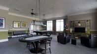This single-floor game room is thoughtfully designed for recreation and social gatherings. The space is anchored by two standout gaming features: an oval poker table with cushioned chairs positioned in the foreground, illuminated by a striking rectangular pendant light suspended from the ceiling, and a full-sized pool table in the background, complete with its own overhead lighting fixture. A ceiling fan adds both functionality and a casual aesthetic to the room.

The walls are painted in a warm olive green, complemented by white crown molding and chair rail detailing that lend a polished, classic character to the space. Large windows along the back wall allow generous natural light to fill the room, framed by floor-length green drapes. The carpeted floor in a subtle grey-blue tone unifies the space and provides comfort underfoot.

A lounge area with plush armchairs is situated near the windows, offering a relaxed seating option away from the gaming tables. An open shelving unit along one wall displays decorative items, while framed artwork adds a finishing touch. Overall, the room balances a recreational focus with refined interior detailing.