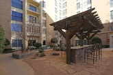 Located at 1001 University Ave in Lubbock, Texas, this outdoor courtyard serves as a central communal space tucked within the footprint of a multi-story brick building complex. The courtyard is thoughtfully designed with a combination of hardscape and softscape elements that create an inviting, functional environment.

At the heart of the space stands a sturdy wooden pergola sheltering a built-in masonry bar counter with tall bar-height seating — an ideal spot for casual gatherings and outdoor socializing. Adjacent to the pergola, a classic iron bench provides additional seating along the walking paths.

The landscaping features a carefully arranged mix of native shrubs, ornamental trees, and decorative boulders set against a gravel and stone ground cover, reflecting a low-maintenance, xeriscape-inspired design well-suited to the West Texas climate. Flagstone pavers define the walkways and patio surfaces, lending a natural, textured aesthetic throughout.

The courtyard is enclosed by the surrounding building structure on multiple sides, offering a sense of privacy and shelter, while wrought iron fencing delineates access points. The multi-story brick facades rising above contribute to the sheltered, enclosed ambiance of the space. Overall, the courtyard presents a well-appointed outdoor retreat that balances functional amenities with attractive, regionally appropriate landscaping.