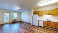 Located at 1710 Pierce St in Lakewood, Colorado, this one-bedroom, one-bathroom unit offers a practical and well-appointed single-floor layout. Warm wood-look flooring runs consistently throughout every room, creating a cohesive and inviting aesthetic. The combined kitchen and living area forms the heart of the unit, with the kitchen featuring warm honey-toned cabinetry, white appliances including a refrigerator, dishwasher, and electric range, ample counter space, and overhead lighting. The open layout allows the kitchen to flow naturally into the living area, which benefits from a private entry door with a window that frames views of surrounding greenery. The bedroom is a comfortable, well-proportioned room with a large window overlooking lush trees, a closet, and a wall-mounted HVAC unit for year-round climate control. A baseboard heater provides additional warmth. The bathroom is thoughtfully designed with wood-look flooring, a warm wood vanity with an integrated sink and mirror, and a walk-in accessible shower/tub area with grab bars. The clean, neutral color palette of soft whites and creams throughout the unit enhances the sense of openness and allows natural light from multiple windows to brighten the space effectively.