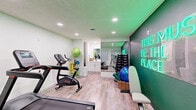 This fitness center in Germantown, Tennessee, is a thoughtfully designed single-room workout space that maximizes every square foot for an efficient and motivating exercise environment. The room features light wood-look flooring throughout, recessed ceiling lighting, and soft green-tinted walls that create a clean, energizing atmosphere.

Cardio equipment is a central focus, with a treadmill, stationary bike, and elliptical machine positioned to allow comfortable use without crowding. A dedicated strength training area lines one wall, where a full rack of dumbbells is neatly organized and reflected by a large floor-to-ceiling mirror that visually expands the space. Wicker baskets at the base of the weight rack hold yoga mats and exercise balls, keeping the area tidy and accessible.

A built-in vanity with white cabinetry and a mirror provides a practical utility station. Natural light filters in through a window fitted with white blinds, complemented by a wall-mounted HVAC unit for year-round climate control.

The standout design feature is a bold, deep green accent wall adorned with a glowing neon sign reading "This Must Be The Place," adding a lively and welcoming focal point to the room. Together, the functional layout and striking aesthetic details make this fitness center an inviting and practical space for daily workouts.