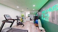 This fitness center in Germantown, Tennessee, is a thoughtfully designed single-room workout space that maximizes every square foot for an efficient and motivating exercise environment. The room features light wood-look flooring throughout, recessed ceiling lighting, and soft green-tinted walls that create a clean, energizing atmosphere.

Cardio equipment is a central focus, with a treadmill, stationary bike, and elliptical machine positioned to allow comfortable use without crowding. A dedicated strength training area lines one wall, where a full rack of dumbbells is neatly organized and reflected by a large floor-to-ceiling mirror that visually expands the space. Wicker baskets at the base of the weight rack hold yoga mats and exercise balls, keeping the area tidy and accessible.

A built-in vanity with white cabinetry and a mirror provides a practical utility station. Natural light filters in through a window fitted with white blinds, complemented by a wall-mounted HVAC unit for year-round climate control.

The standout design feature is a bold, deep green accent wall adorned with a glowing neon sign reading "This Must Be The Place," adding a lively and welcoming focal point to the room. Together, the functional layout and striking aesthetic details make this fitness center an inviting and practical space for daily workouts.