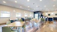 Located at 8855 McGallion Rd in Houston, TX, this single-floor communal dining and kitchen space offers a bright, welcoming environment designed for shared use. The open dining area is furnished with multiple light wood-topped tables paired with a colorful mix of sage green, teal, and navy molded chairs, creating a lively and functional atmosphere. Warm wood-look plank flooring with varied tones runs throughout, adding texture and warmth underfoot. Recessed ceiling lighting is complemented by contemporary white pendant lights that hang above select seating areas, ensuring consistent illumination across the room. A bold deep-blue textured accent wall serves as a striking focal point, adding visual depth and a modern design element to the space. Multiple windows along the perimeter flood the interior with natural light while offering views of the surrounding greenery. The adjacent kitchen area is equally well-equipped, featuring sleek slate-blue lower cabinetry with a clean white countertop, a stainless steel refrigerator, and a microwave. Framed artwork adorns the walls, adding a personalized touch to the functional space. An exit door with standard commercial hardware provides convenient access. Overall, the space balances practicality with a thoughtfully curated, contemporary aesthetic.