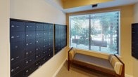 The mail area is a functional and well-appointed space designed to serve residents efficiently. The room is anchored by multiple banks of dark, individually numbered mailboxes mounted directly onto neutral-toned walls, providing organized and secure mail retrieval for a large number of units. The mailboxes are arranged in neat grid formations across two separate wall-mounted panels, ensuring easy access and clear unit identification.

Natural light filters into the space through a large window fitted with white horizontal blinds, offering a pleasant view of mature trees and greenery outside. The window is framed by warm yellow accent walls, adding a welcoming touch to an otherwise utilitarian space. Beneath the window sits a cushioned bench with a wood frame and patterned upholstery, offering a convenient spot for residents while sorting through their mail.

The flooring consists of low-pile carpet in a neutral, multi-toned pattern, contributing to a quiet and comfortable atmosphere. The space connects directly to a hallway corridor, allowing for easy access from other areas of the building. Recessed lighting and a ceiling-mounted smoke detector are also present, reflecting attention to both comfort and safety throughout the area.