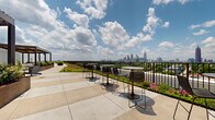 This expansive rooftop terrace offers a thoughtfully designed outdoor retreat elevated above the surrounding cityscape. The terrace is defined by a striking green roof system, where low-growing sedum and flowering plants create a lush, living landscape that borders the perimeter walkways. Corten steel raised planters are arranged throughout the space, filled with a variety of shrubs, flowering plants, and ornamental greenery, adding texture and warmth to the industrial-modern aesthetic.

A slatted wood and steel pergola provides a partially shaded zone, beneath which lounge chairs and a fire bowl create a comfortable area for relaxation. Along the railing edge, high-top bistro tables and bar-height chairs are positioned to take full advantage of the panoramic city skyline views, which stretch across a dense urban horizon under open sky. The large-format concrete pavers underfoot provide a clean, durable surface that connects each zone of the terrace seamlessly.

Access to the terrace is provided through glass-paneled doors set into a corrugated metal facade. Curved bench seating is tucked into quieter garden nooks surrounded by lush plantings, offering more intimate spaces for reflection. The overall design balances greenery, urban views, and functional outdoor furnishings into a cohesive, inviting rooftop environment.