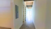 This single-floor space showcases an industrial-modern aesthetic characterized by exposed concrete ceilings and smooth polished concrete floors that lend the interior a clean, contemporary feel. The open corridor leads toward a brightly lit area, suggesting generous natural light penetrating through large windows at the far end — a feature that creates an airy, welcoming ambiance throughout.

The hallway is lined with crisp white walls, offering a neutral canvas that contrasts with the raw, textured ceiling above. A built-in electrical panel is mounted flush on one wall, and an interior door with integrated shelving adds a practical storage element while maintaining the streamlined aesthetic. The exposed structural ceiling elements, including visible beams and utility components, reinforce the industrial character of the space.

The polished concrete flooring flows continuously, unifying the layout and contributing to the sense of openness. The combination of raw materials and clean white finishes gives this space a versatile quality, making it well-suited for use as a studio, creative office, showroom, or professional workspace. Overall, the space presents a functional yet architecturally distinct environment with strong bones and excellent potential for customization.