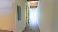 This single-floor space showcases an industrial-modern aesthetic characterized by exposed concrete ceilings and smooth polished concrete floors that lend the interior a clean, contemporary feel. The open corridor leads toward a brightly lit area, suggesting generous natural light penetrating through large windows at the far end — a feature that creates an airy, welcoming ambiance throughout.

The hallway is lined with crisp white walls, offering a neutral canvas that contrasts with the raw, textured ceiling above. A built-in electrical panel is mounted flush on one wall, and an interior door with integrated shelving adds a practical storage element while maintaining the streamlined aesthetic. The exposed structural ceiling elements, including visible beams and utility components, reinforce the industrial character of the space.

The polished concrete flooring flows continuously, unifying the layout and contributing to the sense of openness. The combination of raw materials and clean white finishes gives this space a versatile quality, making it well-suited for use as a studio, creative office, showroom, or professional workspace. Overall, the space presents a functional yet architecturally distinct environment with strong bones and excellent potential for customization.
