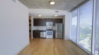 This thoughtfully designed single-floor space combines modern aesthetics with functional living. The open-concept layout seamlessly connects the kitchen and living areas, creating an airy and cohesive environment. The kitchen is well-appointed with dark espresso cabinetry, a subway tile backsplash, and a full suite of stainless steel appliances, including a refrigerator, electric range, and built-in microwave. A stainless steel sink with sleek hardware completes the kitchen's contemporary look. The exposed concrete ceiling adds an industrial character to the space, contrasting beautifully with the warm, light-toned hardwood flooring that extends throughout. Natural light floods the living area through expansive floor-to-ceiling windows, offering views of the surrounding outdoors and creating a bright, welcoming atmosphere. A in-unit stacked washer and dryer is conveniently tucked into a dedicated laundry closet, adding to the practicality of the space. Overall, the unit strikes a balance between urban, industrial design elements and warm, comfortable living — making it an inviting and highly functional space.