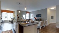This well-appointed single-floor residence showcases an open-concept layout that masterfully connects the kitchen, dining, and living areas into one cohesive and inviting space. The kitchen serves as the heart of the home, featuring a large central island topped with rich granite countertops, an undermount sink, and ample cabinetry with warm wood-toned upper cabinets. Elegant pendant lighting hangs above the island, casting a warm glow over the space and adding a touch of modern sophistication. The kitchen flows naturally into the dining and living areas, where hardwood flooring extends throughout, providing a warm and grounded aesthetic. The living area is bright and airy, benefiting from natural light filtering in through a prominently placed window dressed with a decorative valance. Cool grey walls provide a neutral backdrop that complements both the warm wood tones and the crisp white furnishings. A media console anchors the living space, while the overall layout encourages an easy, comfortable flow between cooking, dining, and relaxing. The open design and thoughtful finishing details make this a functional yet stylish space well-suited for everyday living and entertaining alike.