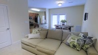 This single-story residence features an open-concept layout that seamlessly connects the living room, dining area, and kitchen into one cohesive, functional space. The living room is anchored by a generously sized sectional sofa in a warm neutral tone, accented with decorative throw pillows in green and white patterns, creating a comfortable and inviting atmosphere. Natural light fills the space through a set of large windows fitted with white blinds, contributing to the bright and airy feel of the interior.

Adjacent to the living area, a compact dining space accommodates a rectangular table with seating for several guests, positioned near the windows to take advantage of the natural light. The kitchen, visible in the background, features rich dark wood cabinetry, stainless steel appliances including a refrigerator, stove, and over-the-range microwave, and a countertop that extends into a breakfast bar with bar stool seating. Light-toned walls throughout the space enhance the sense of openness, while the consistent flooring ties each area together harmoniously. The overall ambiance is clean, casual, and well-organized.