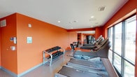 This fitness center is designed to accommodate a well-rounded workout experience within a compact, efficiently organized single-floor layout. The space is immediately distinguished by its bold orange accent walls, which lend an energetic and motivating ambiance throughout. Recessed ceiling lighting provides consistent, even illumination across the entire floor.

Along one side of the room, a row of Precor treadmills is neatly arranged, offering ample cardio training options. A dumbbell rack positioned against the orange wall holds a graduated set of free weights, catering to strength and resistance training needs. Additional cardio and strength equipment, including upright machines and cable systems, are visible toward the rear of the space, maximizing the use of the available floor area.

The flooring consists of a speckled, rubberized surface that provides durability and safety underfoot. A mirrored section toward the back of the room enhances the perception of depth and allows users to monitor their form during workouts. Ventilation is supported by ceiling-mounted HVAC vents. Overall, the space is clean, functional, and thoughtfully arranged to support diverse fitness routines.