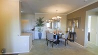 This single-story residence offers a thoughtfully designed open-concept layout that encourages a seamless flow between living spaces. The dining area serves as a focal point, anchored by a round dark-wood table surrounded by upholstered gray chairs and illuminated by a contemporary multi-light chandelier overhead. A large framed mirror on the warm-toned accent wall reflects natural light and adds depth to the room, while decorative framed artwork and a sleek console table with curated accessories complete the refined aesthetic. Soft, neutral-toned carpeting runs throughout, contributing to a warm and comfortable ambiance. A tall potted palm plant introduces a touch of greenery and organic texture to the space. Adjacent to the dining area, a white kitchen counter with a light stone surface is visible, hinting at a well-connected kitchen space designed for both functionality and style. Crown molding along the ceiling adds an architectural detail that elevates the overall finish of the interior. The warm, earthy color palette of the walls, combined with carefully chosen furnishings and decor, creates an inviting atmosphere that is both elegant and livable — ideal for everyday comfort and entertaining alike.