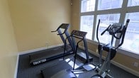 This single-floor fitness room offers a focused cardio workout space equipped with multiple treadmills, including a Trotter model, and an elliptical machine. The room features warm yellow-toned walls that create an inviting atmosphere, complemented by large multi-pane windows that allow natural light to fill the space and provide views to the exterior. The flooring consists of durable, dark carpeting suited for exercise equipment. White baseboards provide a clean contrast to the walls. The layout is efficiently arranged to maximize use of the available space, positioning the cardio machines to take advantage of the natural light from the windows. While compact, the room provides the essential amenities for a cardiovascular workout routine. The large windows not only brighten the space but also offer a connection to the outdoors, making workouts more engaging. Overall, the fitness room maintains a functional and straightforward design focused on delivering a practical exercise environment.