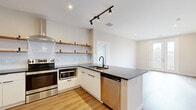 This well-designed single-floor residence showcases a clean, modern aesthetic throughout, characterized by light-toned walls, warm hardwood-style flooring, and an abundance of natural light. The kitchen is a standout feature, offering white flat-front cabinetry with matte black hardware, dark stone countertops, a white subway tile backsplash, and open wood floating shelves. Stainless steel appliances — including a range with a wall-mounted hood, refrigerator, dishwasher, and built-in microwave — complete the space, while a peninsula with an undermount sink and matte black faucet provides additional prep and storage space. The open-plan living area flows seamlessly from the kitchen, bathed in recessed and track lighting. A generously sized bedroom features a ceiling fan, a large window with blinds that invites natural light, and direct access to additional rooms. The bathroom presents a refined look with white cabinetry, dark granite-style countertops, an undermount sink, matte black fixtures, and a sleek rectangular mirror. A walk-in closet offers built-in wire shelving for organized storage. French doors in the living area open to a private balcony, framing scenic views of the surrounding neighborhood and lending the space an airy, elevated feel.